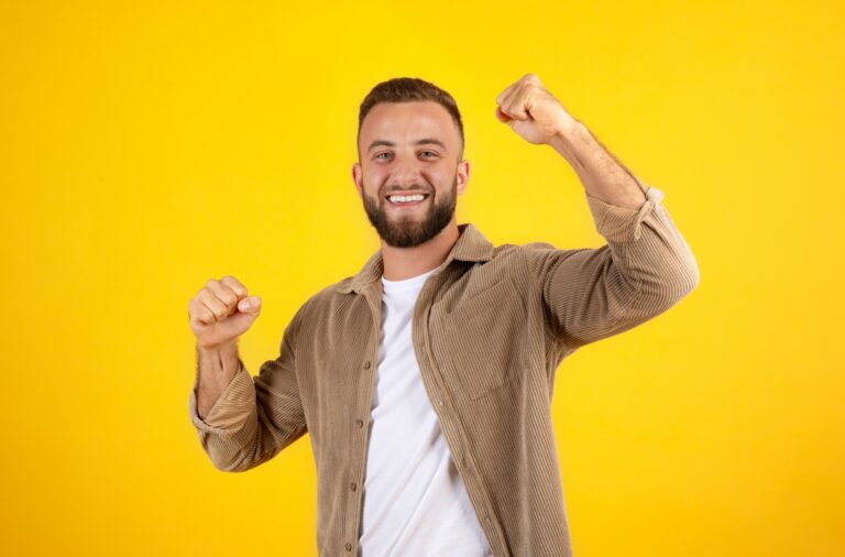 homem-europeu-millennial-feliz-com-barba-em-casual-se-diverte-levantando-os-punhos-para-celebrar-a-vitoria